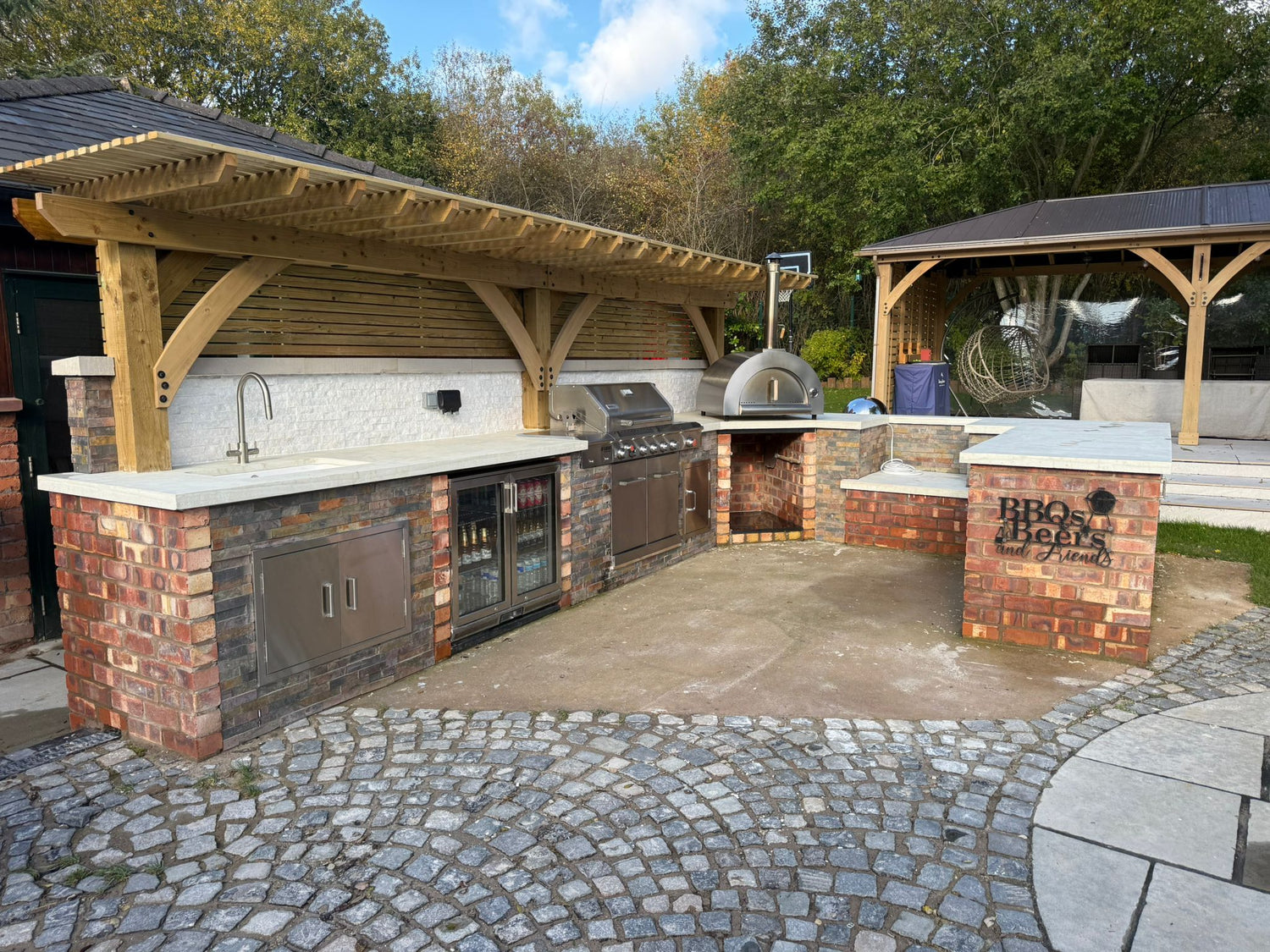 Outdoor kitchen with brick and wood design, featuring a grill and pizza oven, surrounded by trees.
